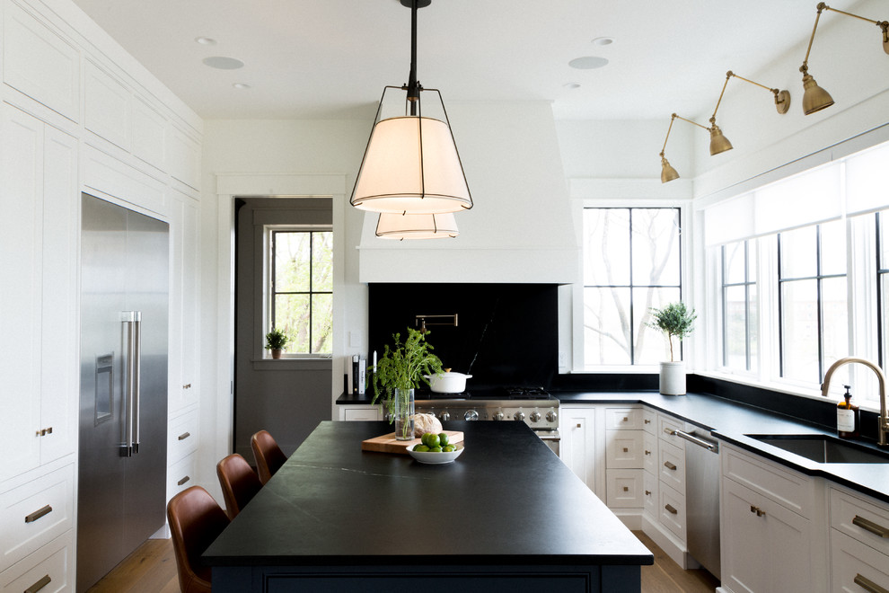 Klassisk inredning av ett stort svart svart kök, med en undermonterad diskho, luckor med infälld panel, vita skåp, bänkskiva i kalksten, svart stänkskydd, stänkskydd i kalk, rostfria vitvaror, ljust trägolv, en köksö och brunt golv