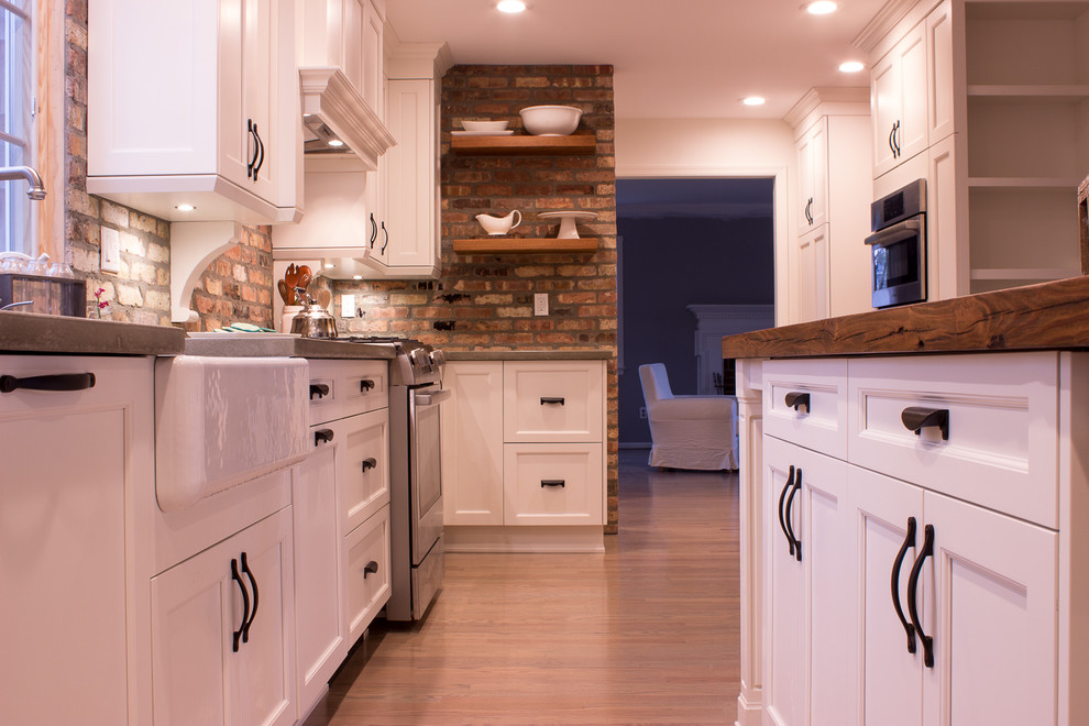 Example of a galley medium tone wood floor and brown floor kitchen design in New York with a farmhouse sink, white cabinets, concrete countertops, red backsplash, brick backsplash, stainless steel appliances and an island