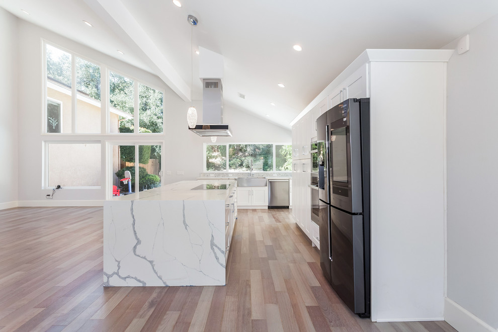 Medium sized modern l-shaped open plan kitchen in Los Angeles with a belfast sink, shaker cabinets, white cabinets, engineered stone countertops, marble splashback, stainless steel appliances, light hardwood flooring, an island and white worktops.