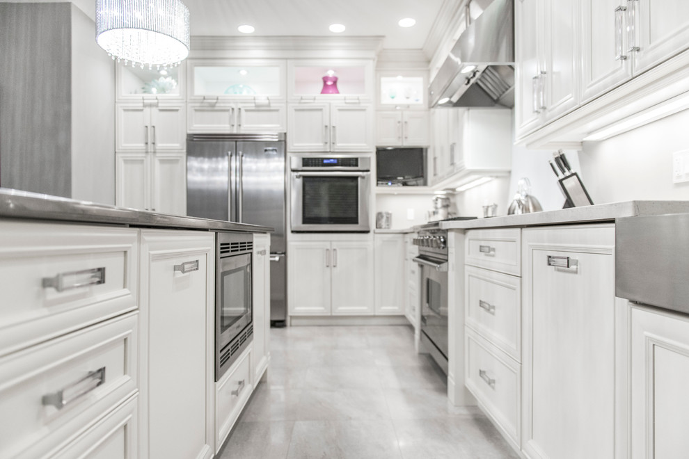 Inredning av ett modernt kök, med en undermonterad diskho, luckor med infälld panel, vita skåp, bänkskiva i rostfritt stål, vitt stänkskydd, rostfria vitvaror, klinkergolv i porslin, en köksö och beiget golv