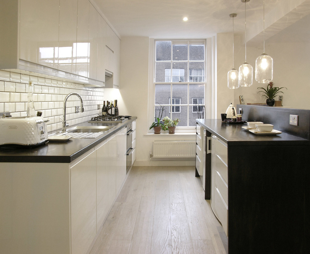 Inspiration for a contemporary galley kitchen in London with a built-in sink, flat-panel cabinets, white splashback, metro tiled splashback, light hardwood flooring and an island.