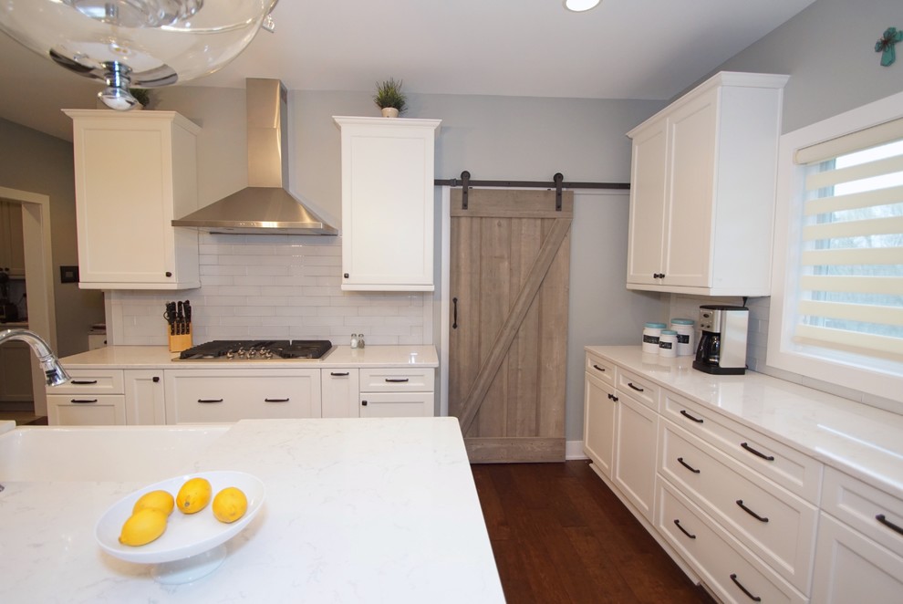Example of a large transitional medium tone wood floor eat-in kitchen design in Omaha with a farmhouse sink, shaker cabinets, white cabinets, quartz countertops, white backsplash, glass tile backsplash, stainless steel appliances and an island