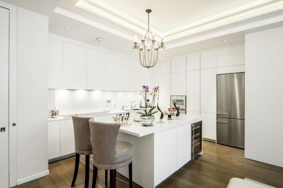 Inspiration for a medium sized contemporary l-shaped kitchen in London with flat-panel cabinets, white cabinets, white splashback, stainless steel appliances, dark hardwood flooring, an island, brown floors and white worktops.