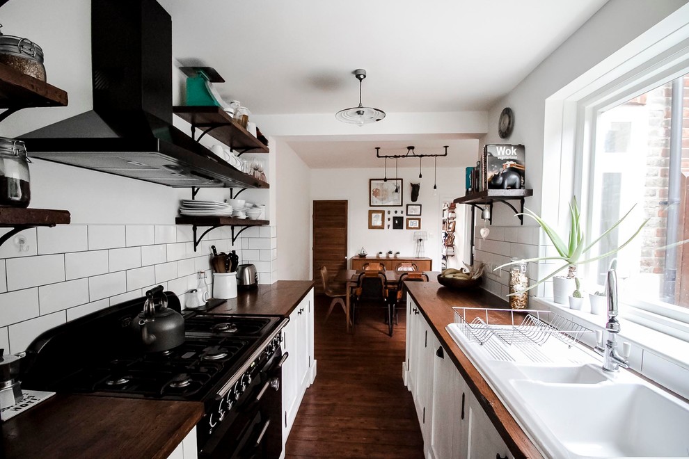 This is an example of a medium sized urban galley open plan kitchen in London with a single-bowl sink, shaker cabinets, white cabinets, wood worktops, white splashback, metro tiled splashback, black appliances, dark hardwood flooring and brown floors.