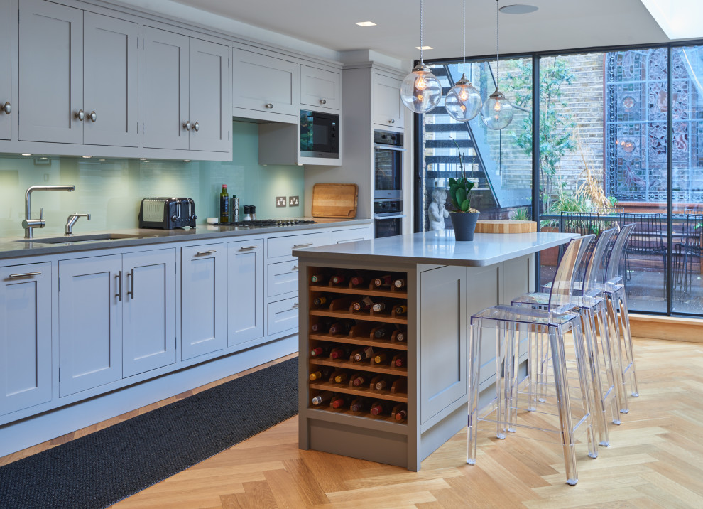 This is an example of a medium sized classic single-wall open plan kitchen in London with a submerged sink, shaker cabinets, grey cabinets, composite countertops, green splashback, glass sheet splashback, stainless steel appliances, light hardwood flooring, an island, brown floors and grey worktops.