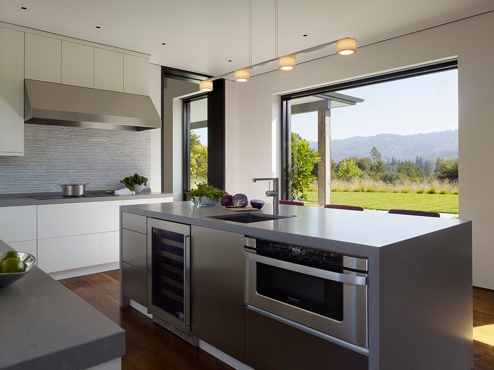 Example of a trendy l-shaped medium tone wood floor kitchen design in San Francisco with flat-panel cabinets, white cabinets, quartz countertops, gray backsplash, stone tile backsplash, stainless steel appliances, an island and a single-bowl sink