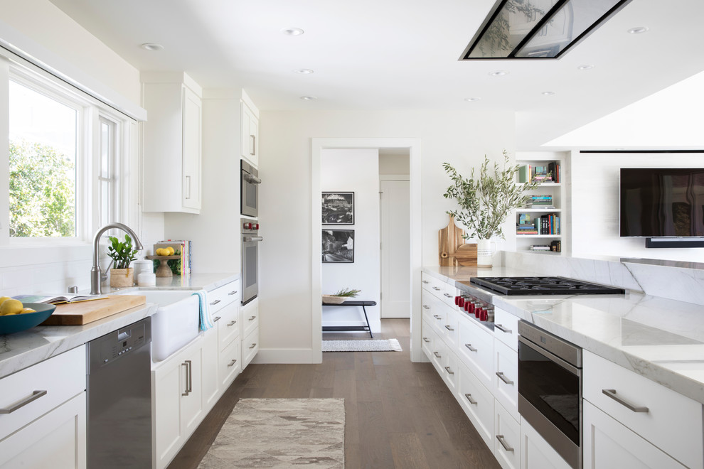 Photo of a medium sized country galley kitchen in San Francisco with a belfast sink, shaker cabinets, white cabinets, engineered stone countertops, white splashback, stainless steel appliances, medium hardwood flooring, an island, brown floors and white worktops.