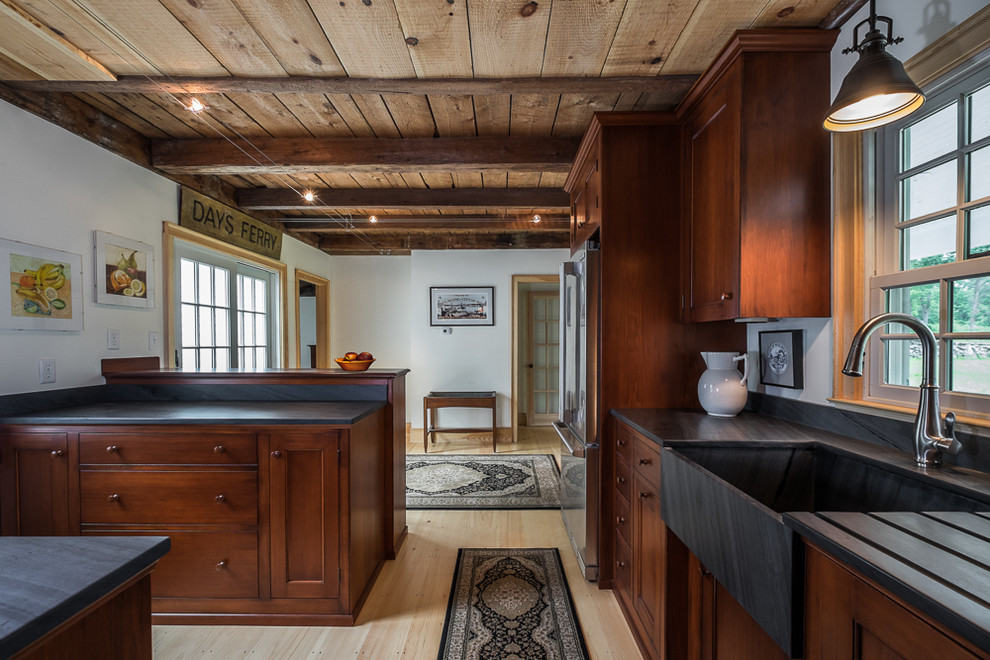 Photo of a small rural l-shaped enclosed kitchen in Portland Maine with a belfast sink, stainless steel appliances, light hardwood flooring, a breakfast bar, recessed-panel cabinets, dark wood cabinets, soapstone worktops and red splashback.