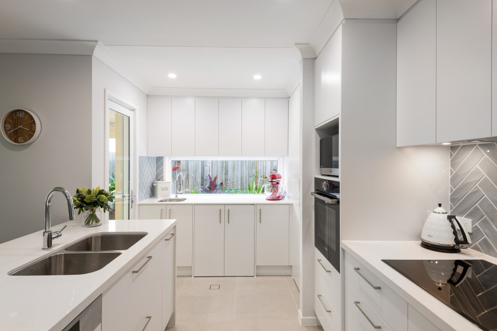 Cette image montre une cuisine minimaliste en L avec un évier 2 bacs, un placard à porte plane, des portes de placard blanches, une crédence grise, un électroménager noir, îlot, un sol gris et un plan de travail blanc.