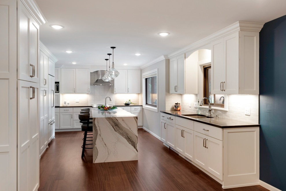 Photo of a large contemporary u-shaped kitchen/diner in Minneapolis with a submerged sink, shaker cabinets, white cabinets, engineered stone countertops, white splashback, porcelain splashback, integrated appliances, dark hardwood flooring, an island, brown floors and grey worktops.