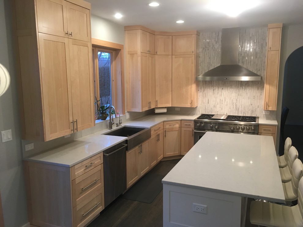 Medium sized traditional l-shaped kitchen in Chicago with a belfast sink, glass tiled splashback, white appliances, dark hardwood flooring, brown floors, shaker cabinets, light wood cabinets, laminate countertops, white splashback and an island.