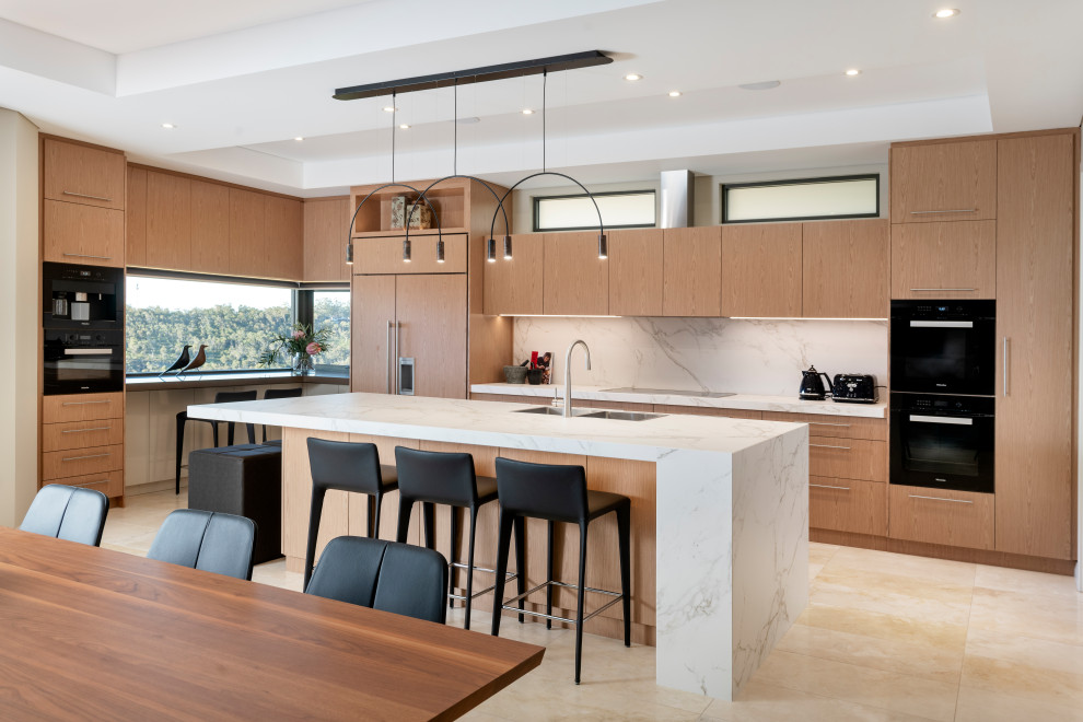 Large contemporary l-shaped open plan kitchen in Perth with a double-bowl sink, recessed-panel cabinets, light wood cabinets, white splashback, black appliances, travertine flooring, an island, beige floors and white worktops.