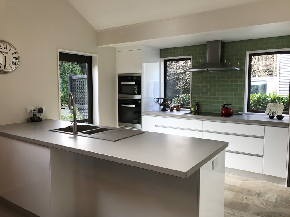 This is an example of a large contemporary galley open plan kitchen in Christchurch with a double-bowl sink, white cabinets, engineered stone countertops, green splashback, glass tiled splashback, stainless steel appliances, porcelain flooring, a breakfast bar and multi-coloured floors.