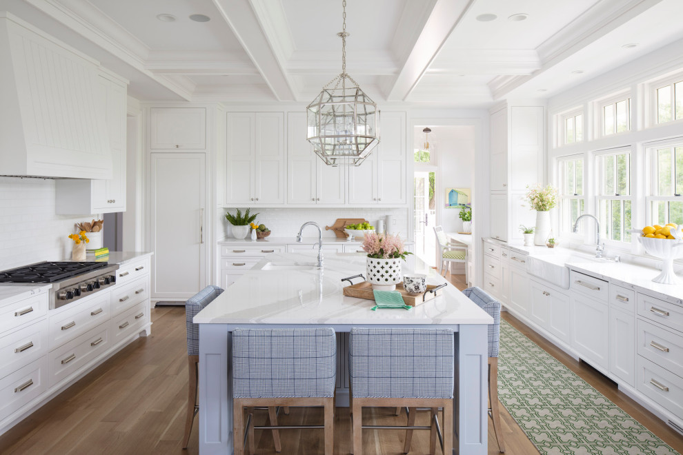Inredning av ett klassiskt stort vit vitt kök, med en rustik diskho, luckor med infälld panel, vita skåp, bänkskiva i kvarts, vitt stänkskydd, stänkskydd i tunnelbanekakel, integrerade vitvaror, mellanmörkt trägolv, en köksö och brunt golv