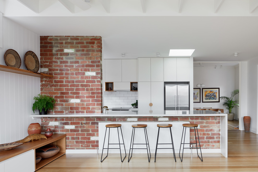Photo of a contemporary open plan kitchen in Melbourne with flat-panel cabinets, white cabinets, white splashback, metro tiled splashback, stainless steel appliances, light hardwood flooring, a breakfast bar, beige floors and white worktops.