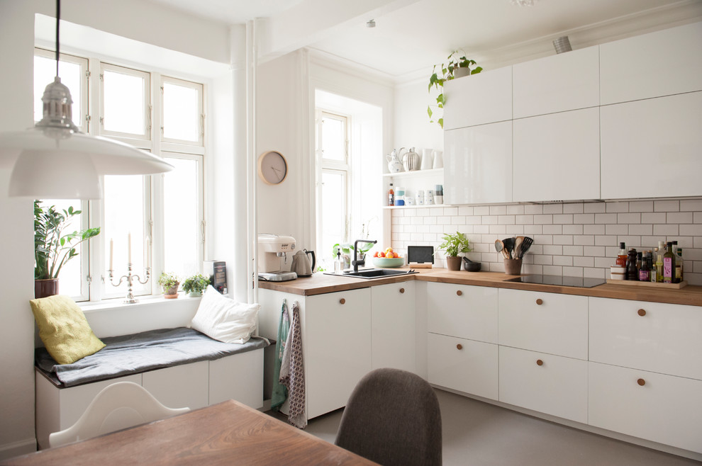 На фото: угловая кухня в скандинавском стиле с обеденным столом, плоскими фасадами, белыми фасадами, деревянной столешницей, белым фартуком, фартуком из плитки кабанчик, накладной мойкой и серым полом без острова с