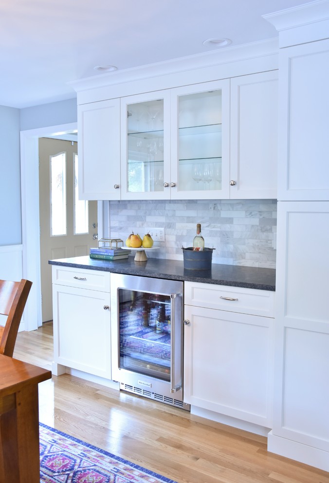 This is an example of a traditional kitchen/diner in Boston with a belfast sink, recessed-panel cabinets, white cabinets, granite worktops, marble splashback, stainless steel appliances, light hardwood flooring and an island.