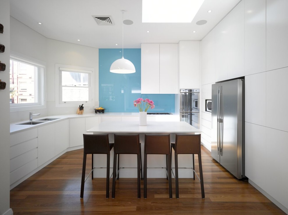 Foto på ett mellanstort funkis kök, med en undermonterad diskho, släta luckor, vita skåp, bänkskiva i kvarts, blått stänkskydd, stänkskydd i sten, rostfria vitvaror, mörkt trägolv och en köksö