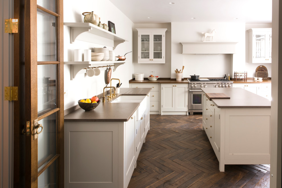 Inspiration for a large traditional enclosed kitchen in London with shaker cabinets, white cabinets, stainless steel appliances, dark hardwood flooring, an island, brown floors, brown worktops and a belfast sink.