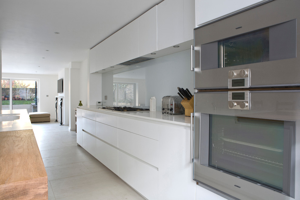 Design ideas for a large contemporary kitchen/diner in London with flat-panel cabinets, white cabinets, glass sheet splashback, stainless steel appliances, an island, a submerged sink, composite countertops, white splashback and porcelain flooring.