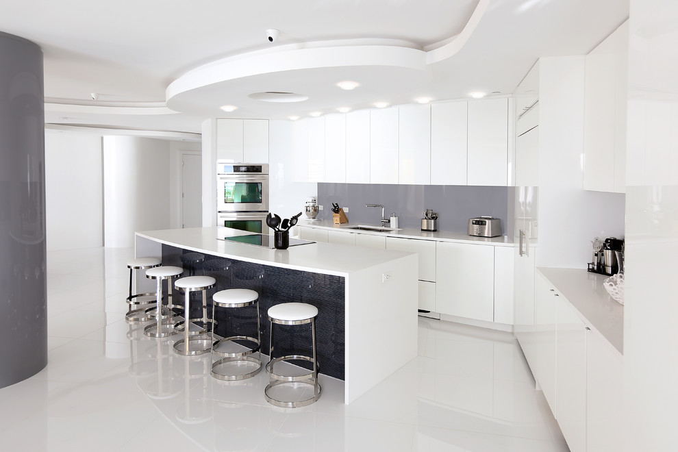 Moderne Küche in L-Form mit Unterbauwaschbecken, flächenbündigen Schrankfronten, weißen Schränken, Küchenrückwand in Grau, Glasrückwand, Küchengeräten aus Edelstahl und Kücheninsel in Sonstige