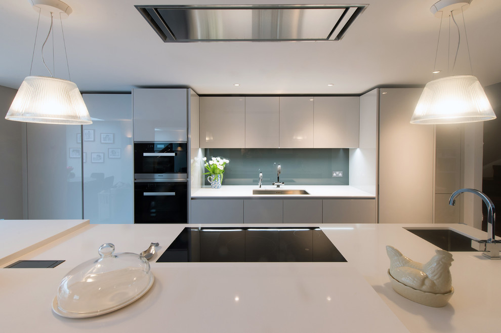 Photo of a small contemporary open plan kitchen in London with an integrated sink, flat-panel cabinets, grey cabinets, composite countertops, grey splashback, glass sheet splashback, black appliances and an island.
