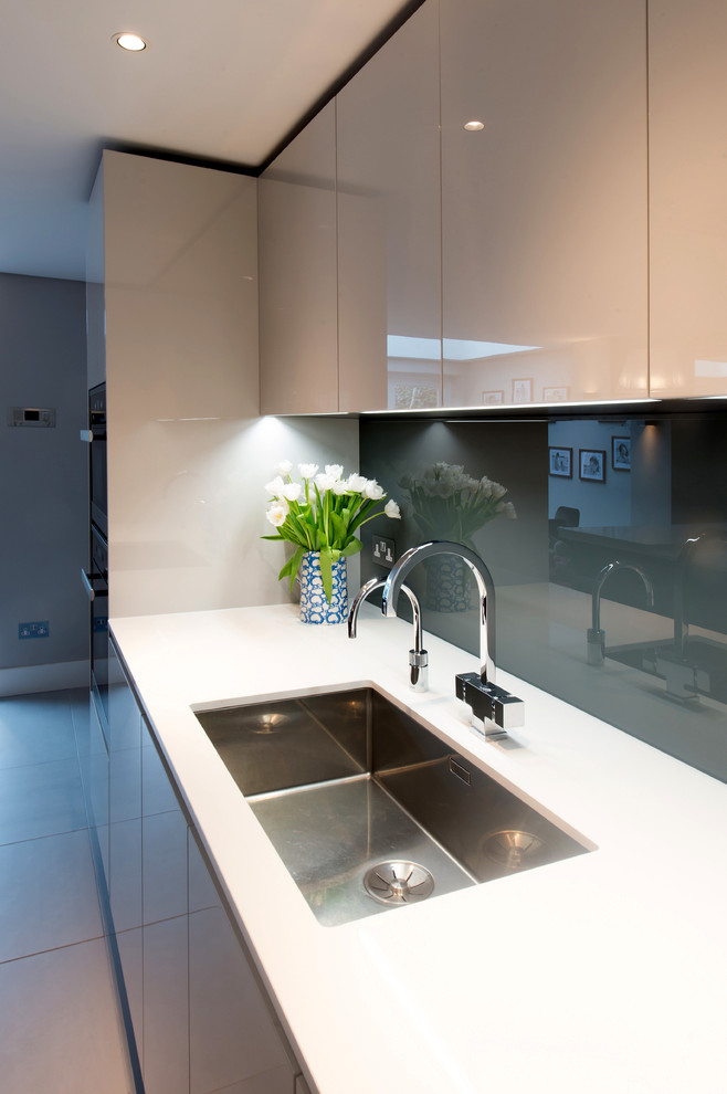 Example of a small trendy open concept kitchen design in London with an integrated sink, flat-panel cabinets, gray cabinets, solid surface countertops, gray backsplash, glass sheet backsplash, black appliances and an island