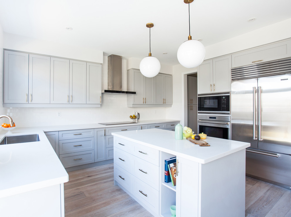 Photo of a medium sized contemporary l-shaped kitchen in Toronto with a submerged sink, shaker cabinets, white cabinets, composite countertops, white splashback, stainless steel appliances, light hardwood flooring and an island.