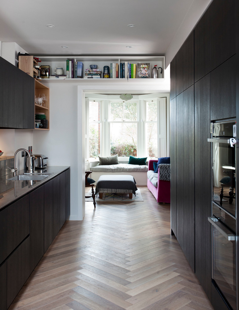 Exempel på ett mellanstort modernt kök, med en integrerad diskho, släta luckor, skåp i mörkt trä, bänkskiva i rostfritt stål, stänkskydd med metallisk yta, glaspanel som stänkskydd, integrerade vitvaror, mellanmörkt trägolv, en halv köksö och flerfärgat golv