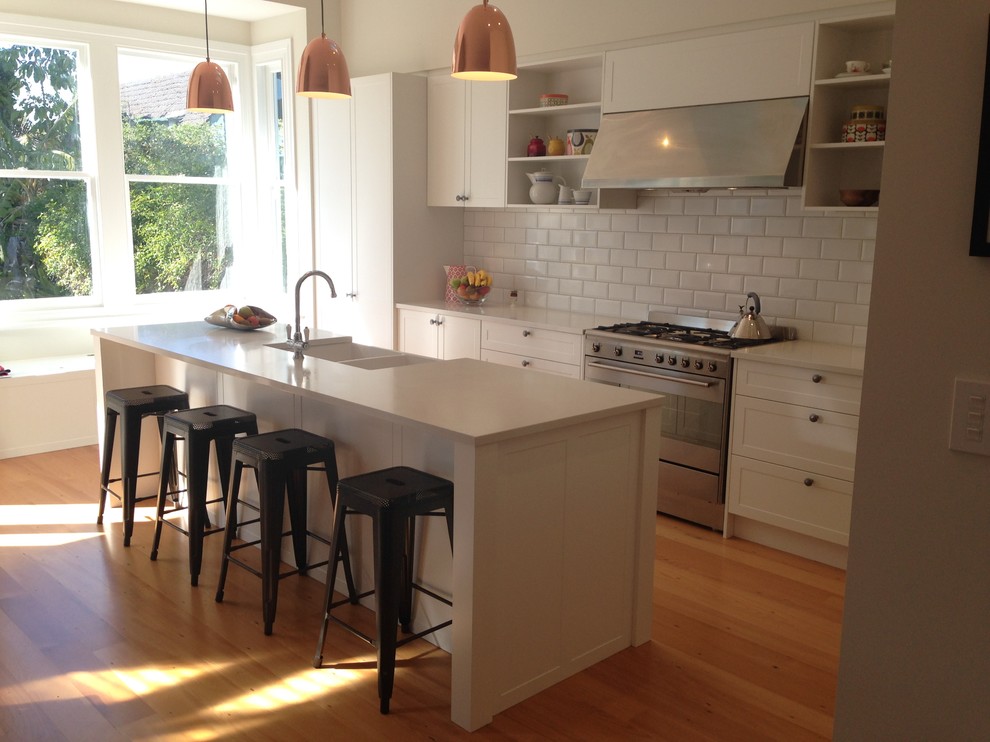Photo of a large traditional l-shaped open plan kitchen in Auckland with a belfast sink, recessed-panel cabinets, white cabinets, engineered stone countertops, white splashback, metro tiled splashback, stainless steel appliances, medium hardwood flooring and an island.