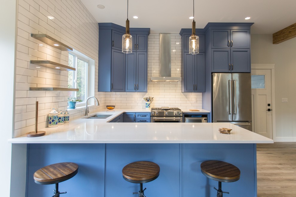 Foto på ett mellanstort lantligt vit u-kök, med en undermonterad diskho, luckor med infälld panel, blå skåp, bänkskiva i kvartsit, vitt stänkskydd, stänkskydd i tunnelbanekakel, rostfria vitvaror, ljust trägolv, en halv köksö och beiget golv