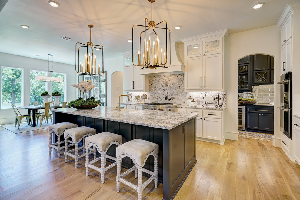 Inspiration for a transitional l-shaped light wood floor and brown floor kitchen remodel in Oklahoma City with a farmhouse sink, recessed-panel cabinets, white cabinets, gray backsplash, stone slab backsplash, stainless steel appliances, an island and gray countertops