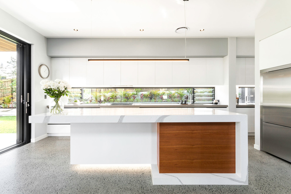 Bild på ett stort funkis vit vitt kök, med släta luckor, vita skåp, fönster som stänkskydd, rostfria vitvaror, en köksö, grått golv, en nedsänkt diskho, bänkskiva i kvarts och betonggolv