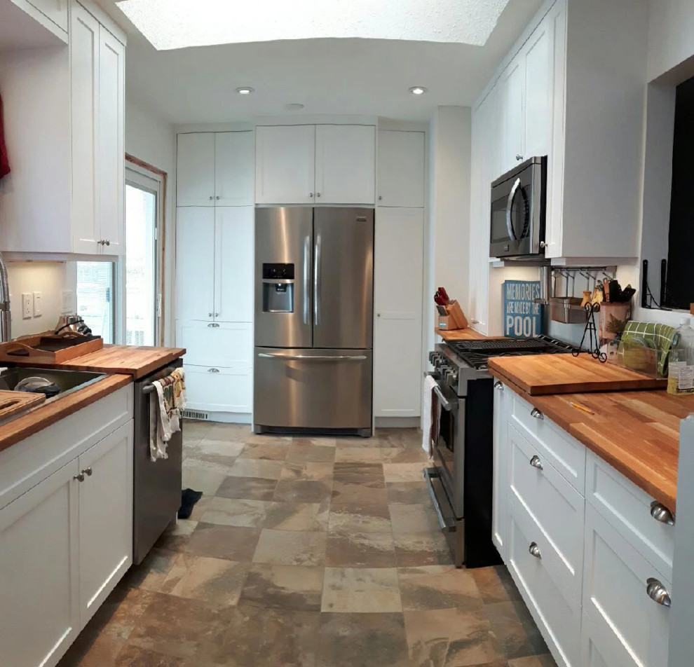 На фото: отдельная, параллельная кухня среднего размера в стиле кантри с накладной мойкой, фасадами в стиле шейкер, белыми фасадами, деревянной столешницей и техникой из нержавеющей стали
