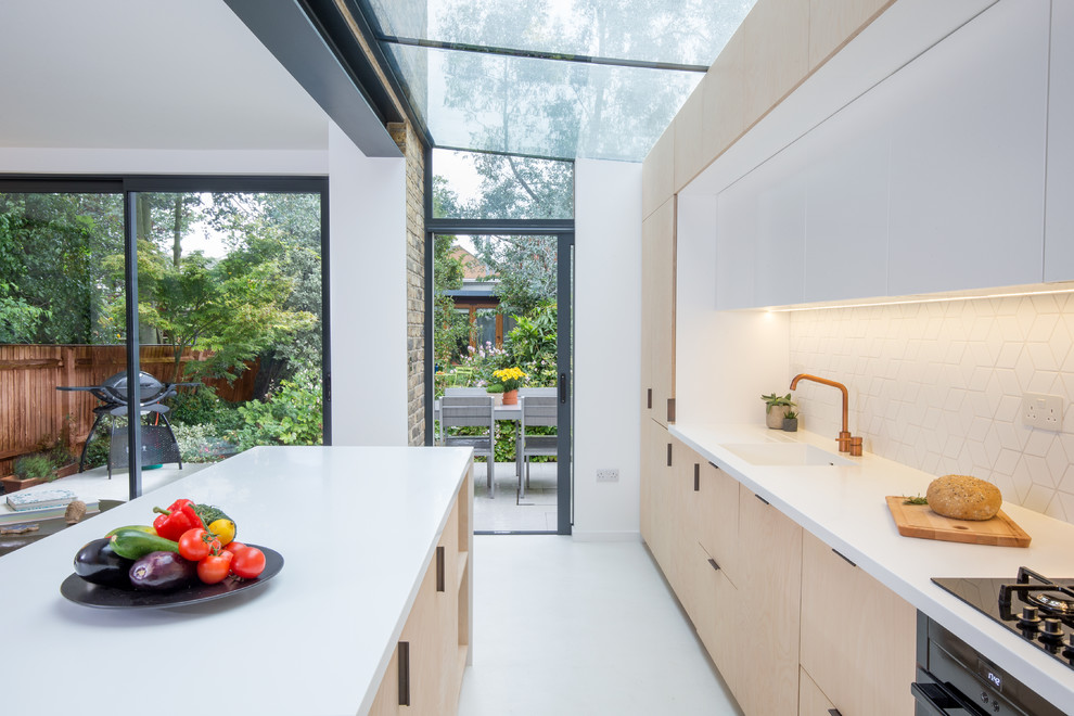 Идея дизайна: маленькая прямая кухня-гостиная в стиле модернизм с монолитной мойкой, плоскими фасадами, светлыми деревянными фасадами, белым фартуком, фартуком из керамической плитки, черной техникой, бетонным полом, островом и белым полом для на участке и в саду