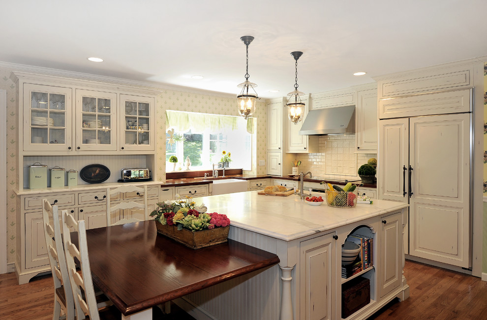 Mid-sized elegant l-shaped medium tone wood floor and brown floor kitchen photo in New York with a farmhouse sink, raised-panel cabinets, white cabinets, marble countertops, white backsplash, subway tile backsplash, paneled appliances and an island