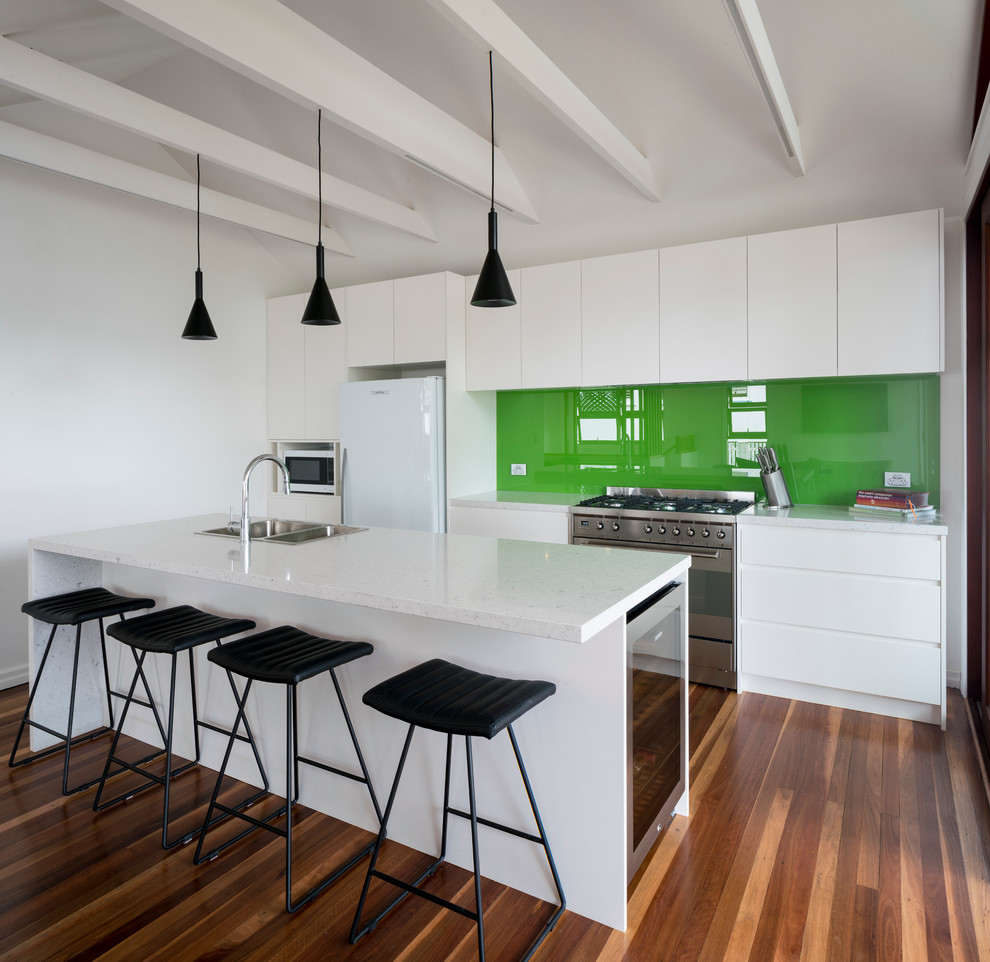 Design ideas for a modern galley open plan kitchen in Brisbane with a double-bowl sink, white cabinets, composite countertops, green splashback, glass sheet splashback, stainless steel appliances, medium hardwood flooring and an island.