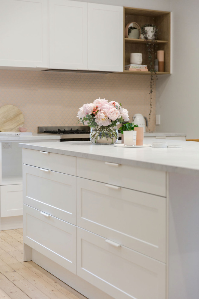Photo of a small scandi l-shaped kitchen/diner in Melbourne with a submerged sink, recessed-panel cabinets, white cabinets, engineered stone countertops, pink splashback, mosaic tiled splashback, black appliances, medium hardwood flooring, an island and white worktops.