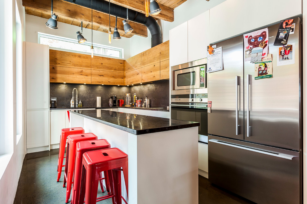 Foto på ett industriellt l-kök, med släta luckor, skåp i mellenmörkt trä, svart stänkskydd, rostfria vitvaror, betonggolv, en köksö och grått golv