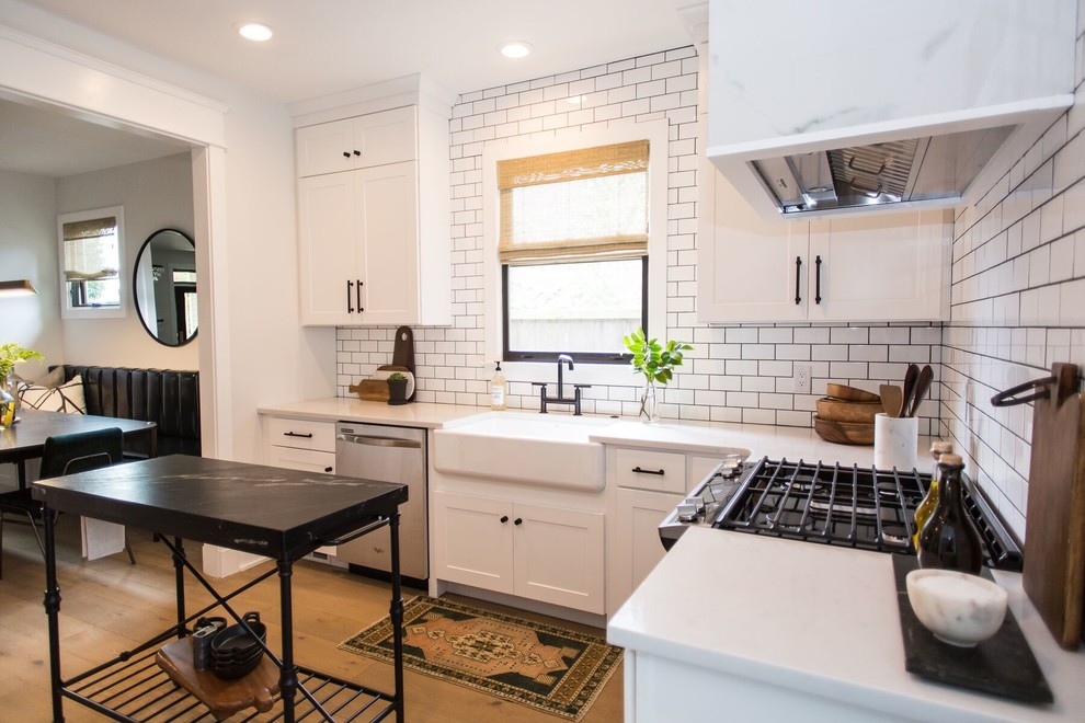 This is an example of a classic l-shaped kitchen/diner in Nashville with a belfast sink, recessed-panel cabinets, white cabinets, white splashback, metro tiled splashback, stainless steel appliances and light hardwood flooring.