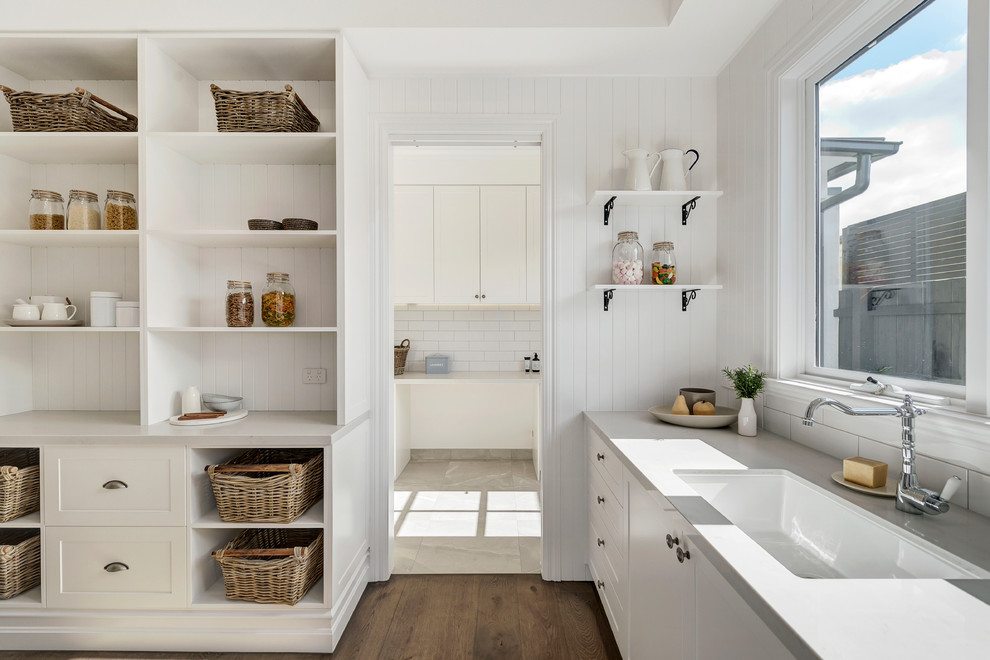 Example of a beach style l-shaped medium tone wood floor and brown floor kitchen design in Melbourne with an undermount sink, shaker cabinets, white cabinets, white backsplash and gray countertops