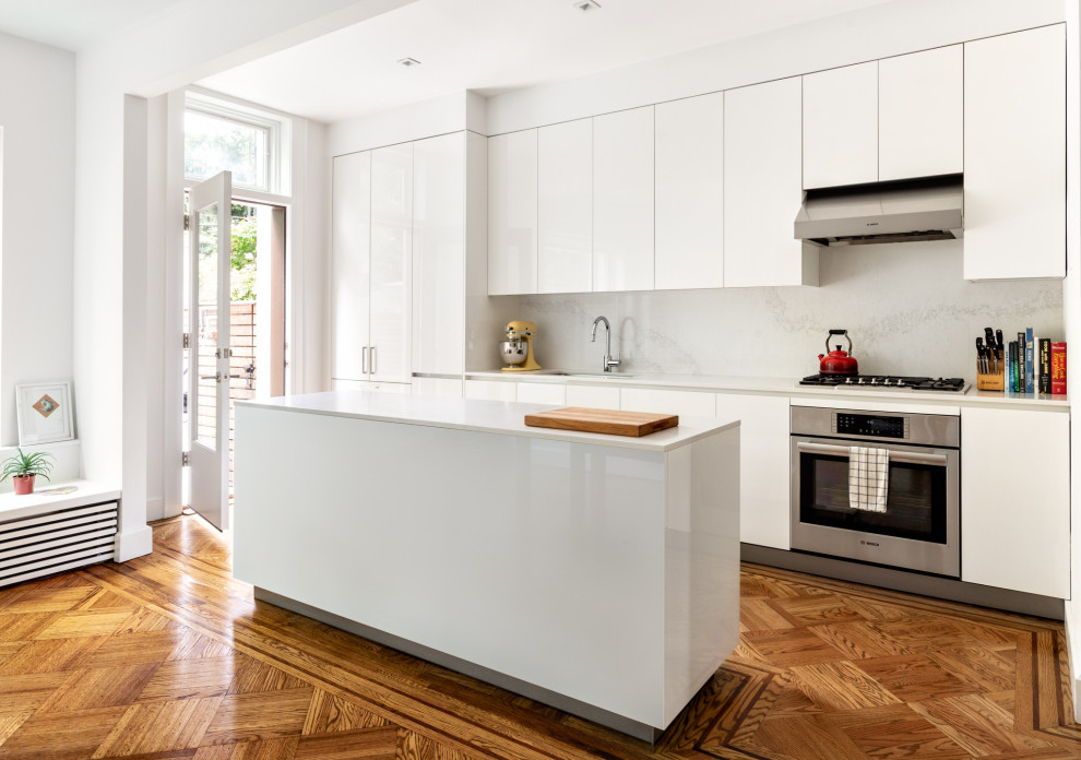Inspiration for a contemporary galley kitchen in New York with a submerged sink, flat-panel cabinets, white cabinets, white splashback, stone slab splashback, stainless steel appliances, medium hardwood flooring, an island, brown floors and white worktops.