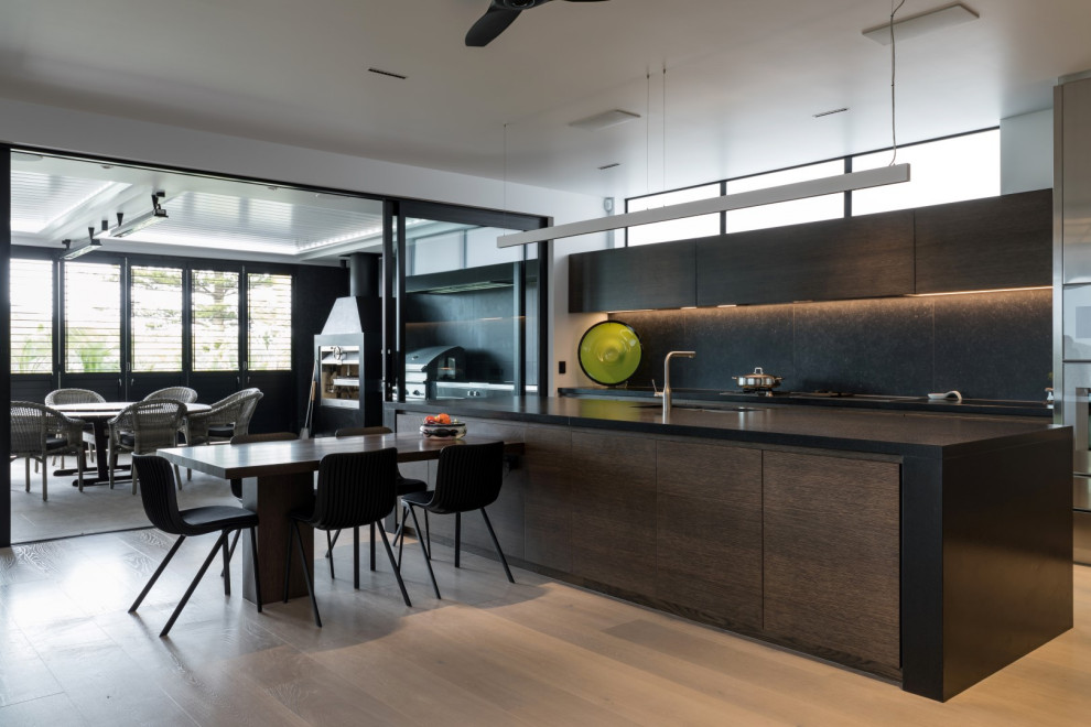 Photo of a large modern galley kitchen/diner in Other with a double-bowl sink, beaded cabinets, dark wood cabinets, composite countertops, black splashback, cement tile splashback, stainless steel appliances, light hardwood flooring, an island, beige floors and black worktops.