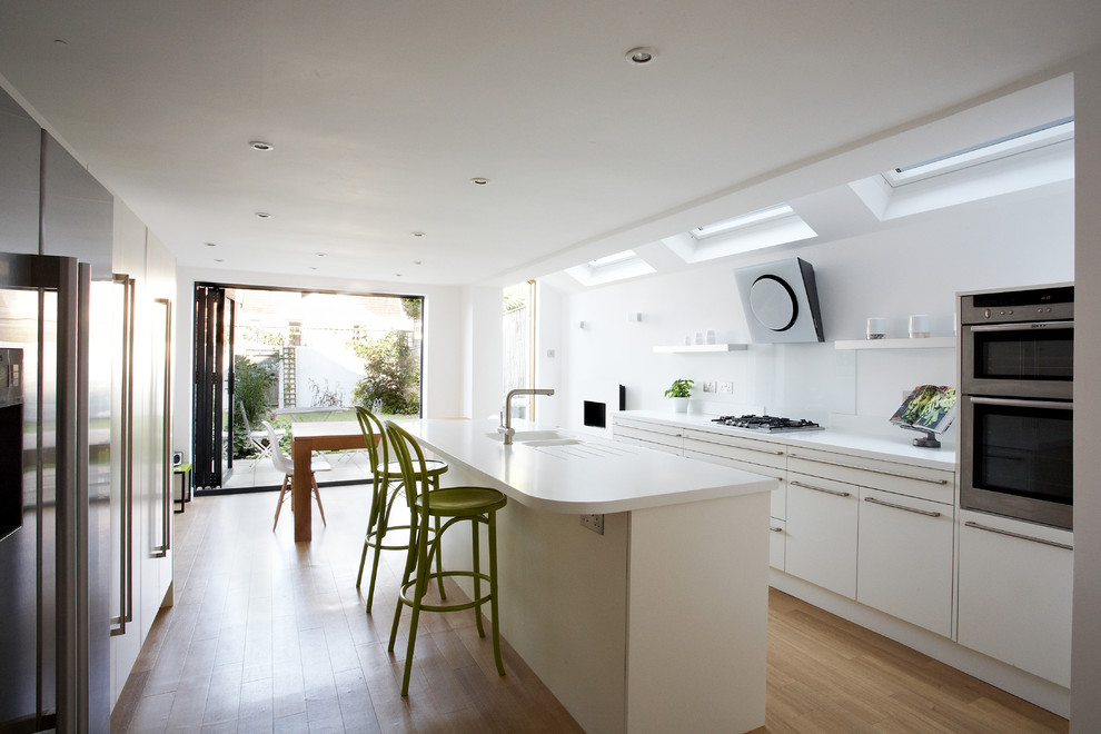 Moderne Wohnküche mit integriertem Waschbecken, flächenbündigen Schrankfronten, weißen Schränken, Küchenrückwand in Weiß, Küchengeräten aus Edelstahl, hellem Holzboden und Kücheninsel in Sonstige