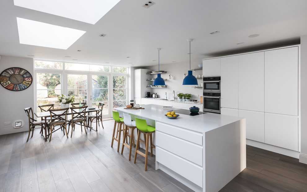 Offene, Große, Zweizeilige Moderne Küche mit flächenbündigen Schrankfronten, weißen Schränken, Küchengeräten aus Edelstahl, hellem Holzboden und Kücheninsel in London