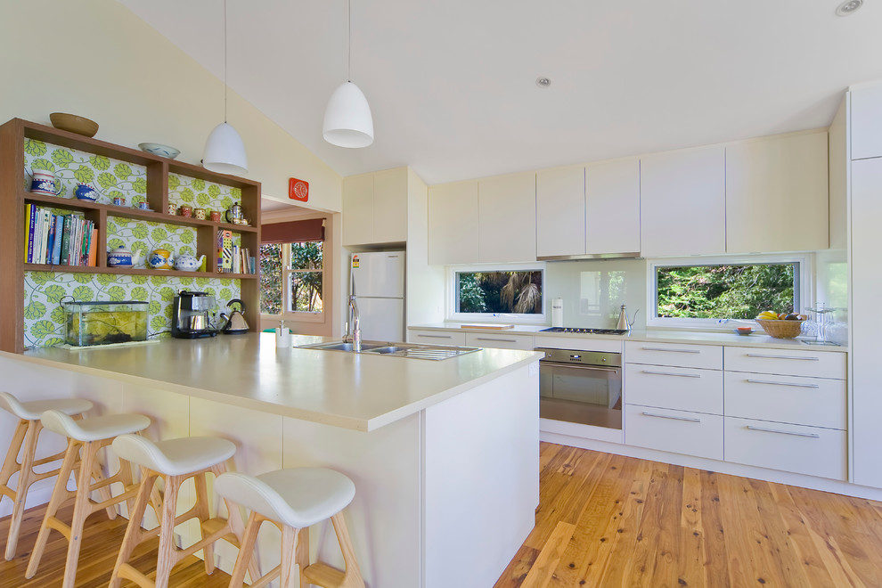 Photo of a medium sized contemporary galley open plan kitchen in Sydney with a double-bowl sink, flat-panel cabinets, white cabinets, engineered stone countertops, white splashback, glass sheet splashback, stainless steel appliances, light hardwood flooring and an island.