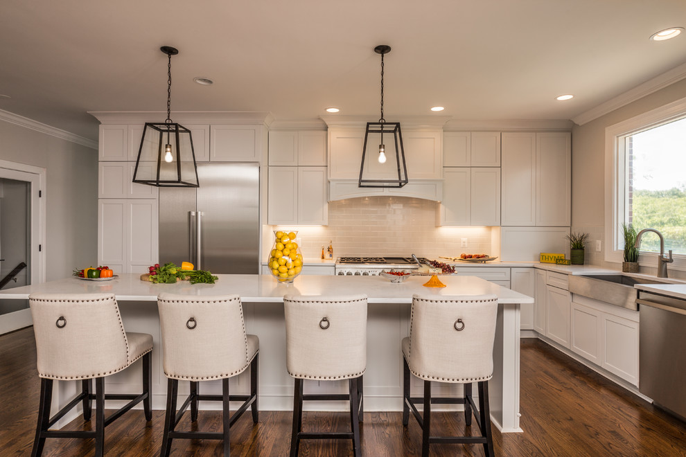 Large classic l-shaped kitchen/diner in DC Metro with a belfast sink, shaker cabinets, white cabinets, beige splashback, stainless steel appliances, dark hardwood flooring, an island, engineered stone countertops and porcelain splashback.