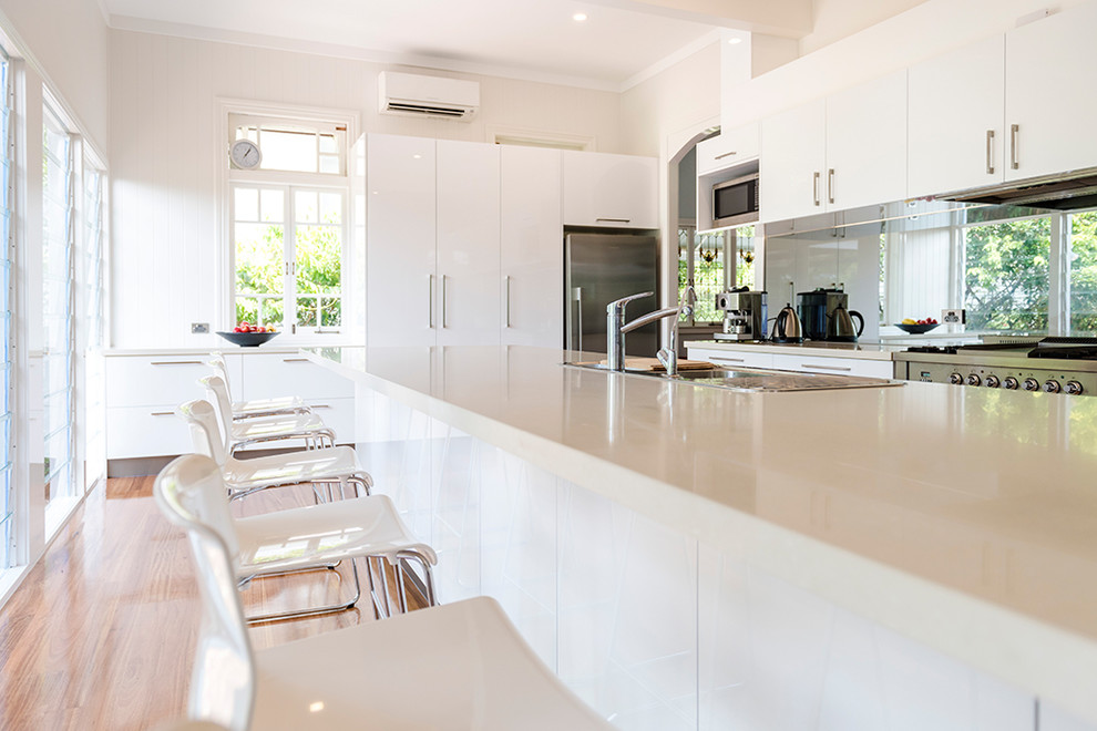 Example of a mid-sized urban u-shaped medium tone wood floor and brown floor eat-in kitchen design in Brisbane with a drop-in sink, flat-panel cabinets, white cabinets, glass sheet backsplash, an island, white countertops, quartz countertops and stainless steel appliances