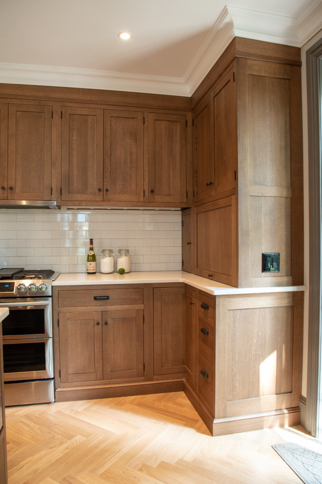 Exemple d'une cuisine américaine craftsman en L et bois brun de taille moyenne avec un évier encastré, un placard à porte shaker, un plan de travail en quartz modifié, une crédence blanche, une crédence en céramique, un électroménager en acier inoxydable, parquet clair, îlot et un plan de travail blanc.