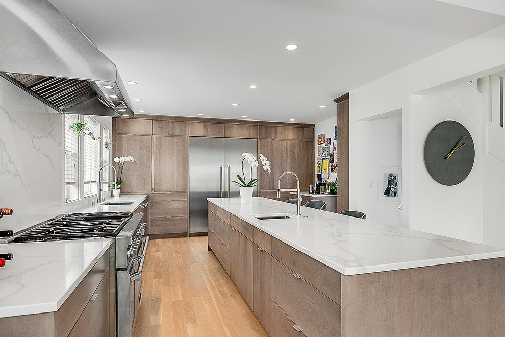 Photo of a large contemporary l-shaped open plan kitchen in Seattle with a submerged sink, flat-panel cabinets, medium wood cabinets, engineered stone countertops, white splashback, stone slab splashback, stainless steel appliances, light hardwood flooring, an island, beige floors and white worktops.